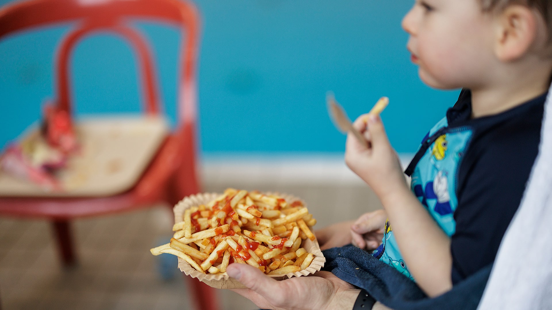 Ett barn sitter med en tallrik pommes frites i handen bredvid en bassäng i en simhall.